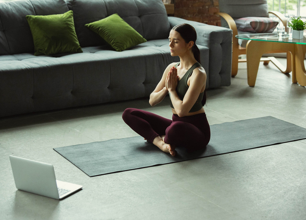 Yoga online junge Frau sitzt im Schneidersitz auf ihrer Yogamatte mit einem geöffneten Laptop vor ihr